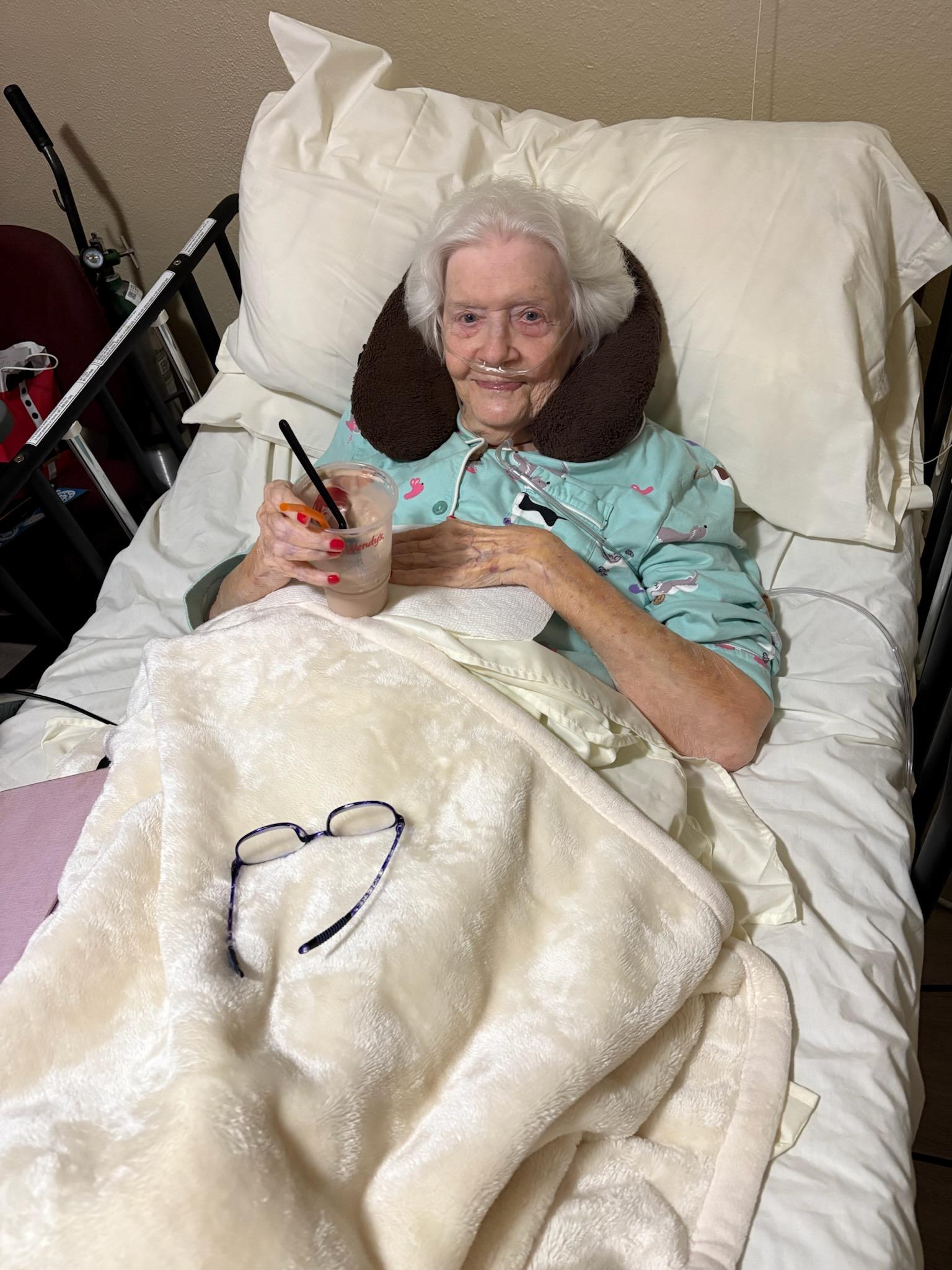 An elderly woman relaxes in bed, sipping a drink and smiling warmly with cozy blankets around her.