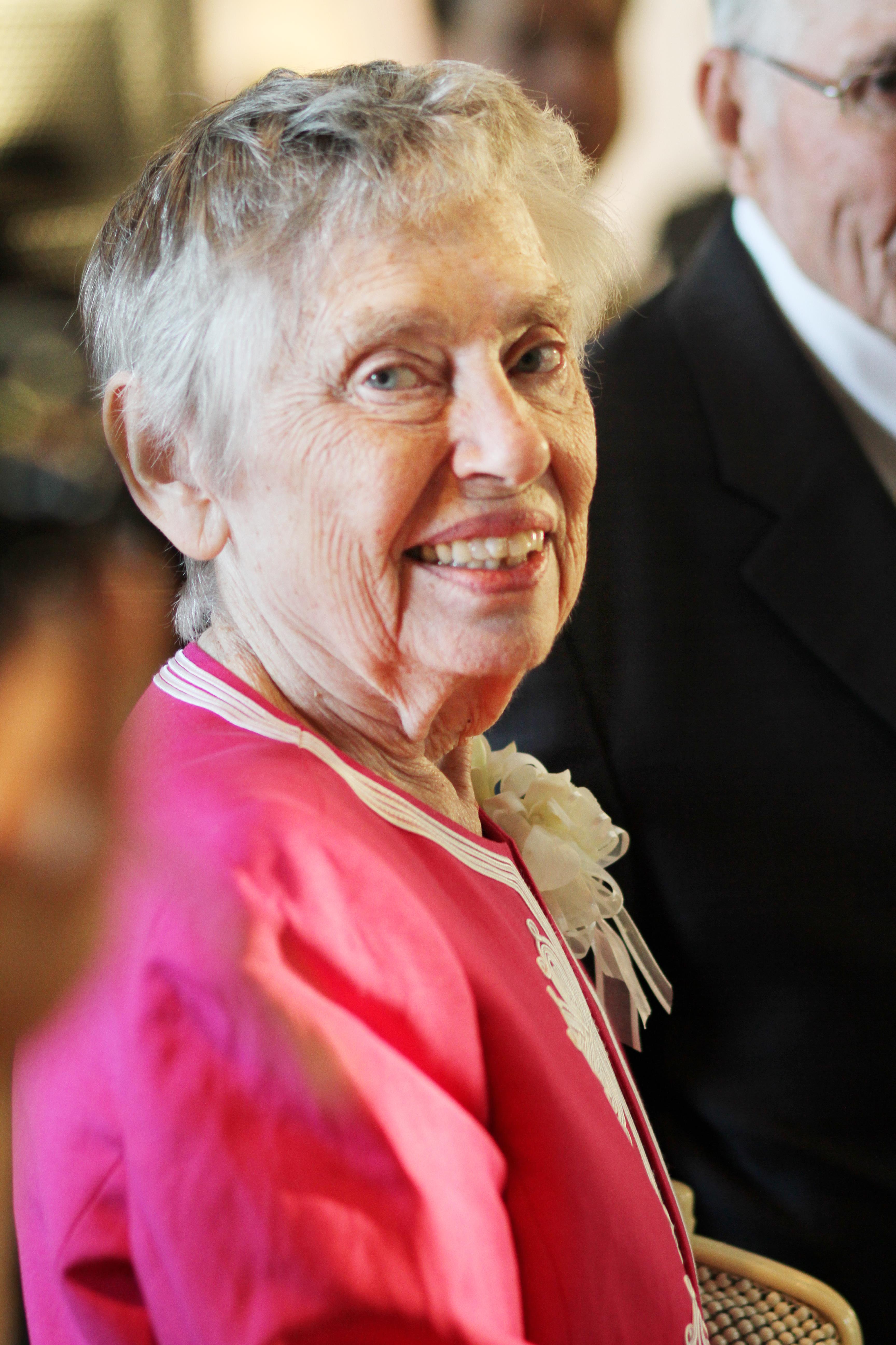 Elderly woman dressed in a vibrant pink outfit smiles warmly at a gathering filled with attendees.