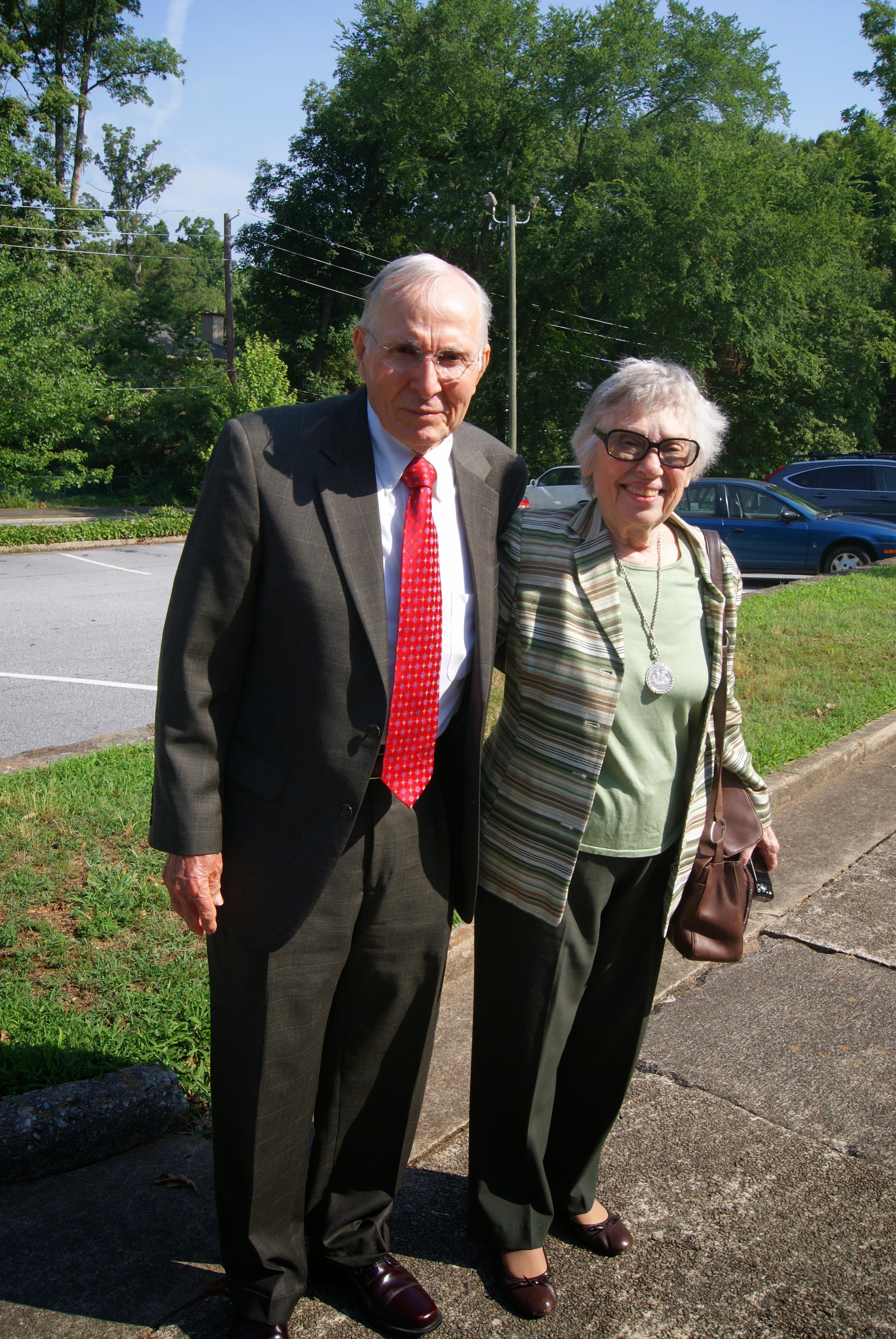 An elderly couple in formal wear smiles together under the sun in a park.