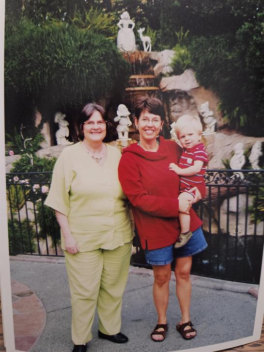 Two smiling women stand in a garden with a child, surrounded by greenery and decorative statues.