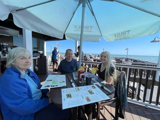 Three people relax and share a meal at an outdoor table overlooking the ocean.