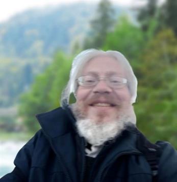 A man with glasses and a beard smiles happily while outdoors by the water.