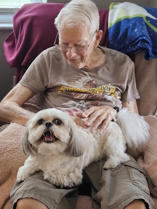 A senior man sits comfortably in a chair, smiling as he pets his happy dog on his lap.
