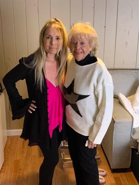 Two women stand side by side, smiling, showcasing their fashionable outfits in a comfortable room.