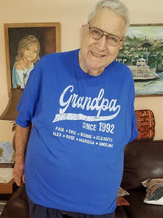 Cheerful older man in a blue grandparent shirt stands beside a sofa in a warm home setting.