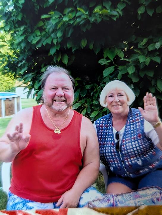 Two cheerful individuals wave at the camera while seated outdoors under a tree.