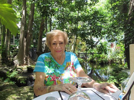 A joyful elderly woman relaxes at a table surrounded by tropical plants and a pond.