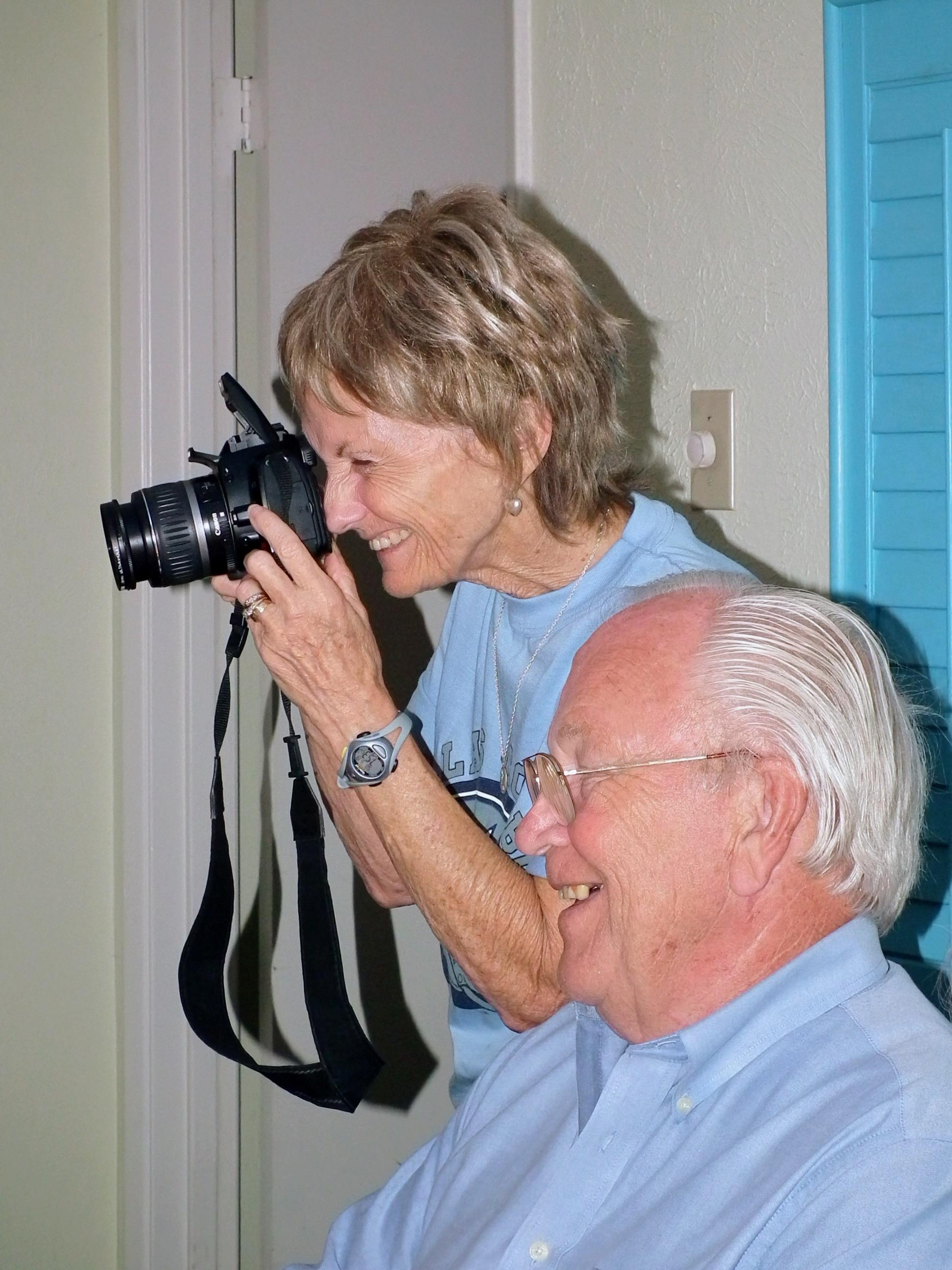 Senior woman happily photographs elderly man in a cheerful indoor setting, sharing laughter.