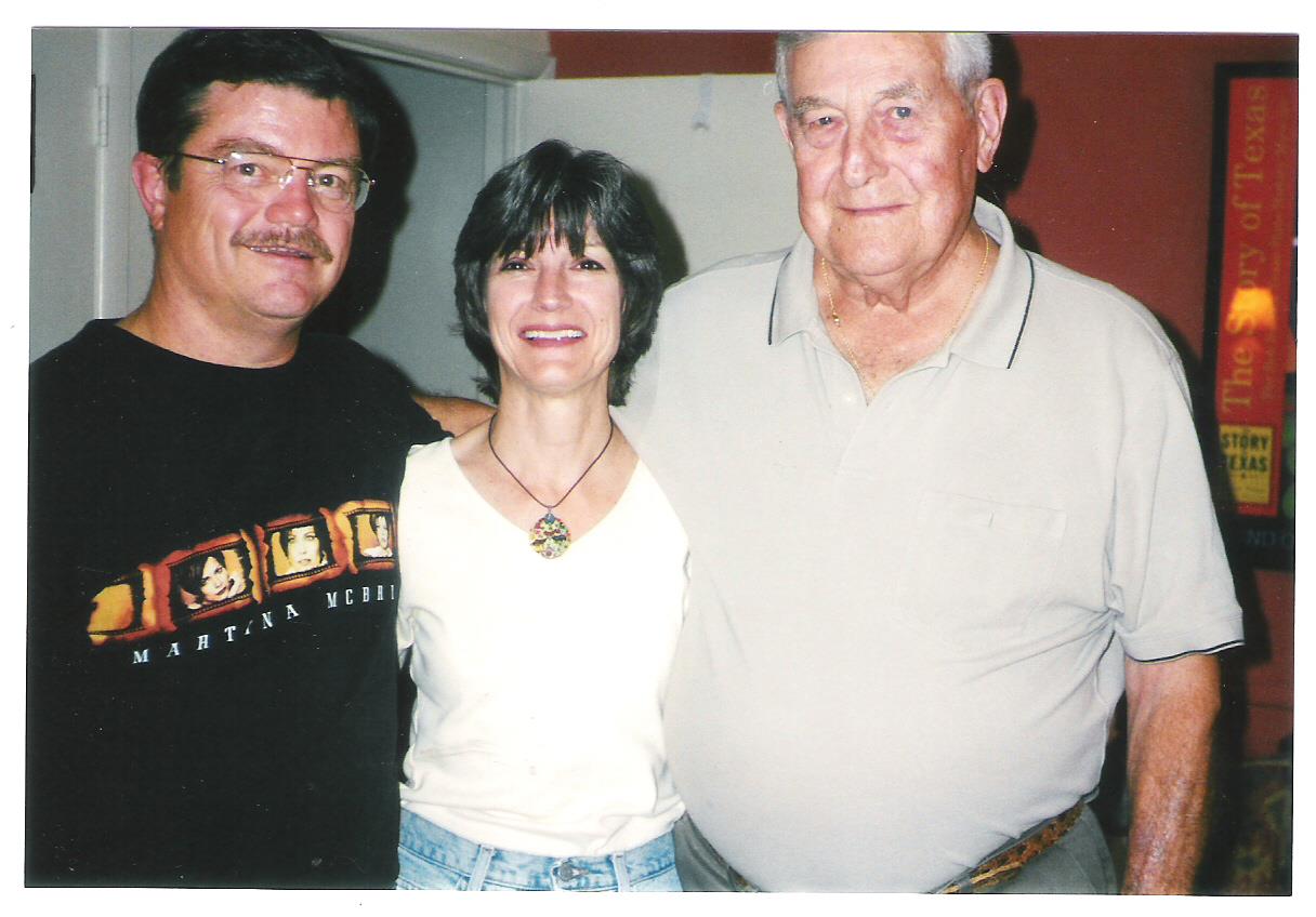 Three people stand close together, smiling happily in a relaxed indoor environment.