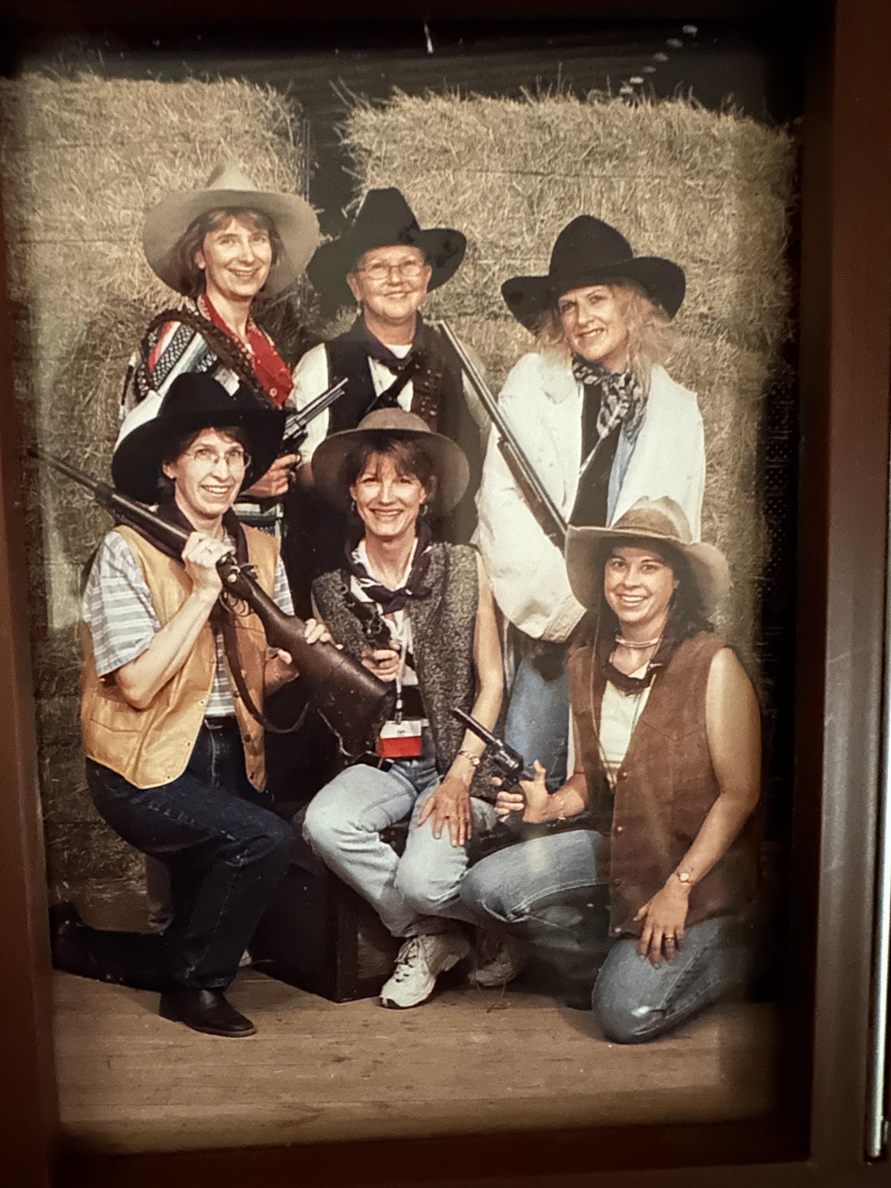 Women celebrate a western theme event, dressed in cowboy attire, holding rifles and smiling.