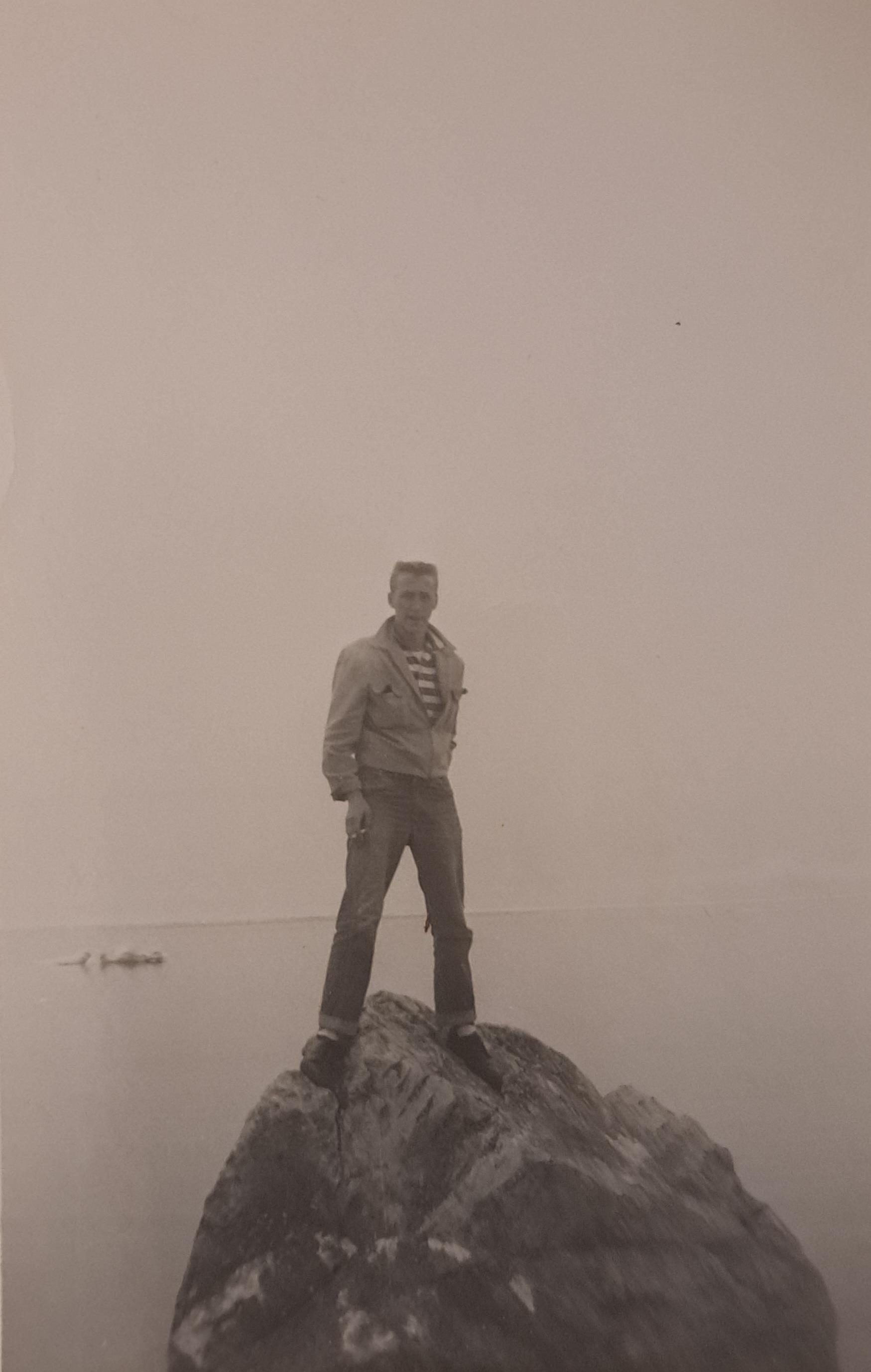 A person stands on a large rock near the ocean with waves gently lapping around them.