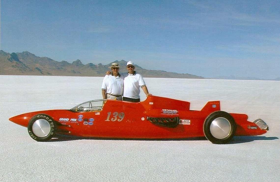 Two individuals in a red racing car engage in speed trials on a vast salt flat landscape.