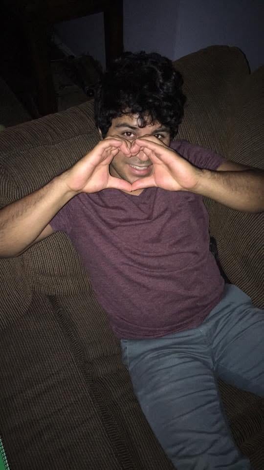 A young man sits on a couch, forming a heart shape with his hands and smiling happily.