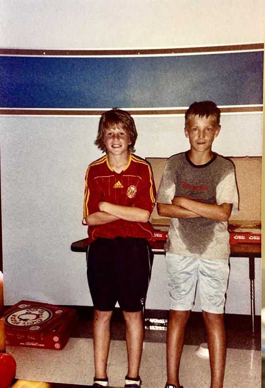 Two boys stand with arms crossed, smiling, after spending time together indoors in summer.