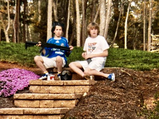 Two boys relax on stone steps in a garden, one holding an airsoft gun, enjoying snacks together.