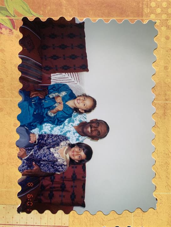 Two joyful children in traditional attire sit beside a smiling adult in a comfortable living space.