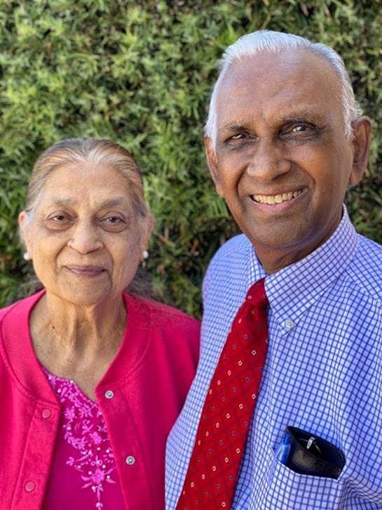 A happy elderly couple smiles at the camera, dressed in colorful attire in their garden.