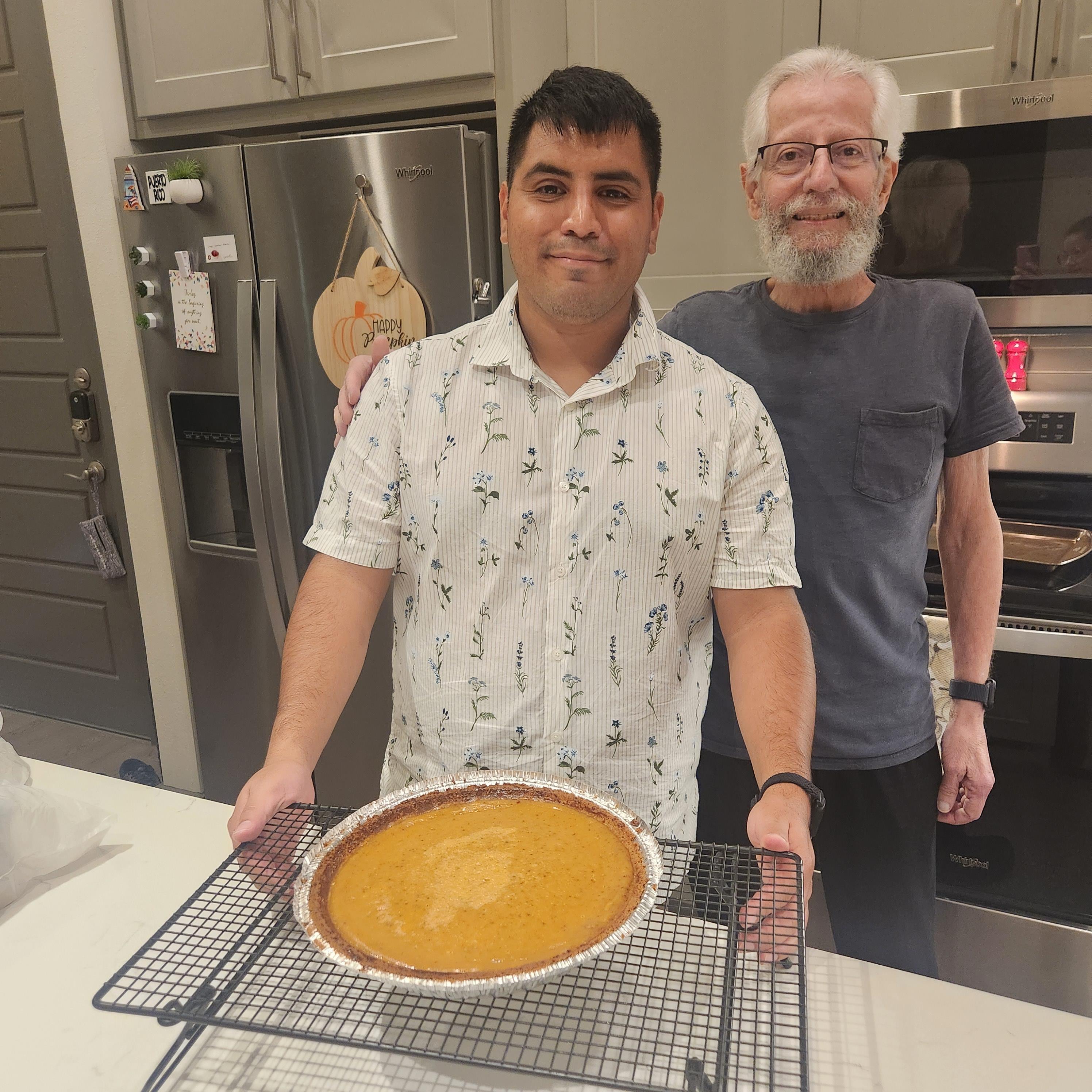 A man holds a warm pumpkin pie while another stands beside him in a stylish kitchen.