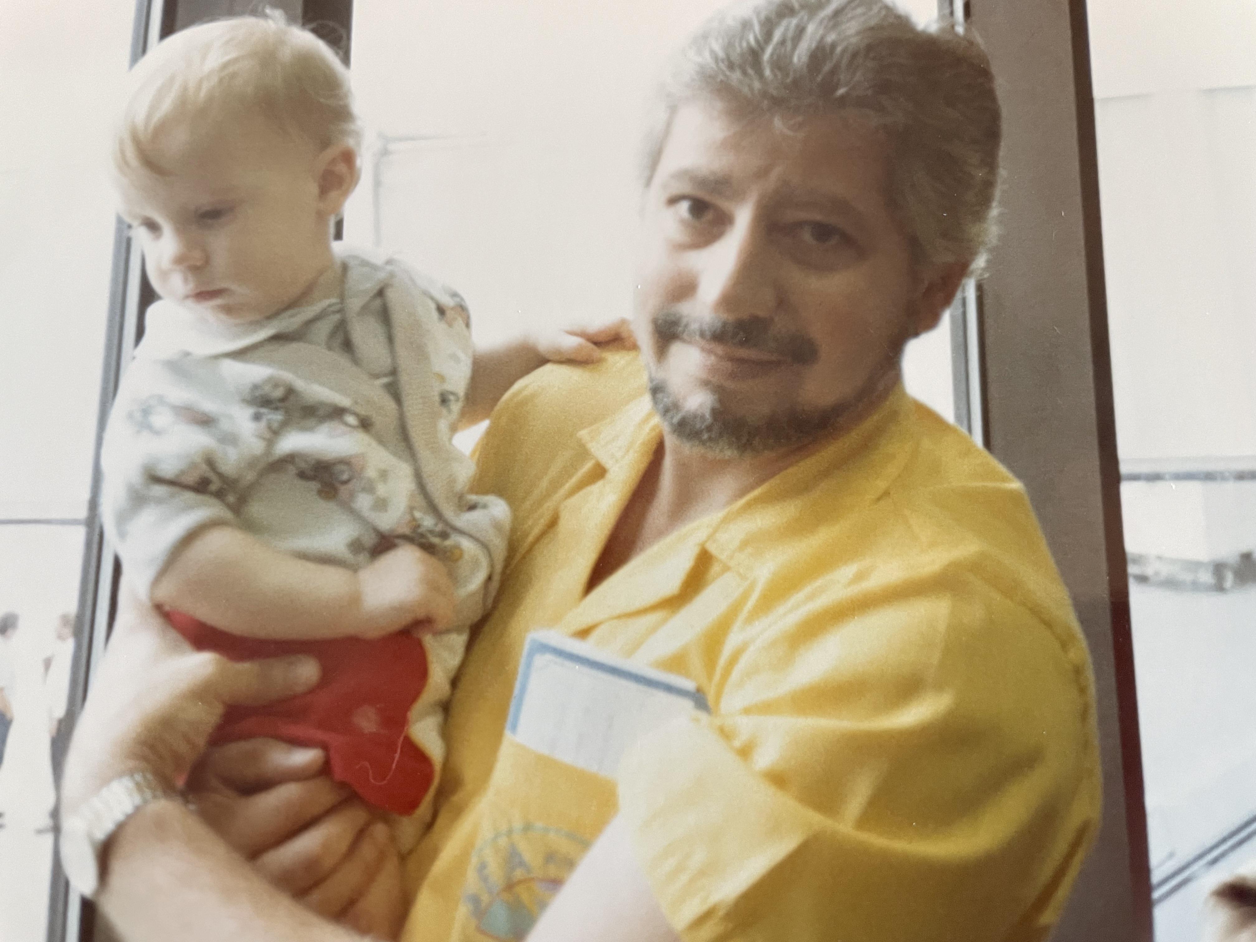 A man holds a young child with a gentle smile while indoors close to a window.