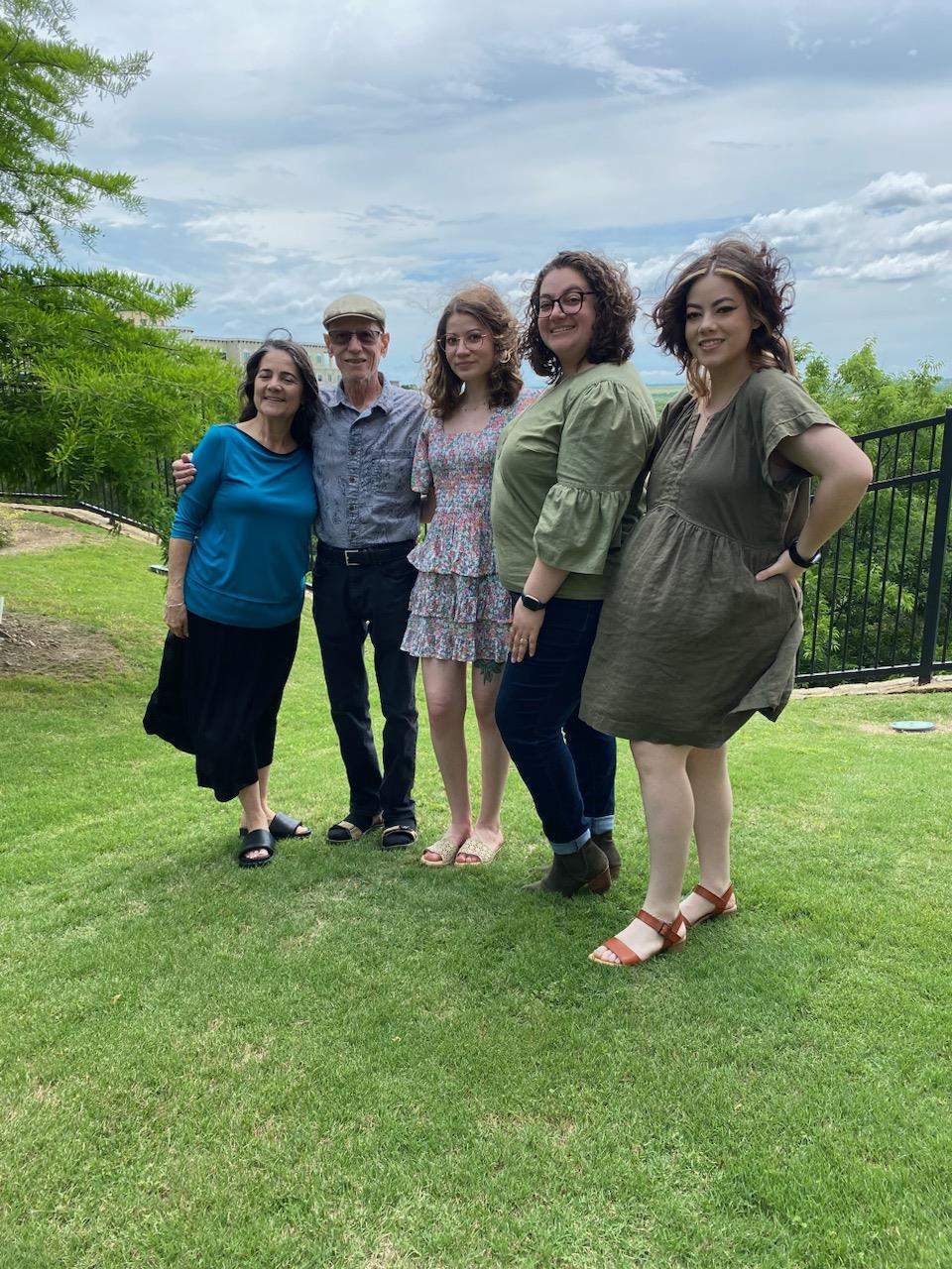 Five family members stand together on green grass with trees and clouds in the background.