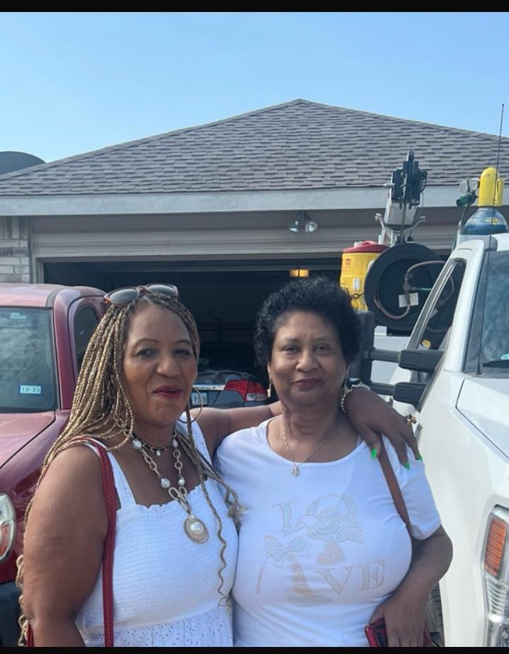 Two smiling women stand closely together in front of a garage, enjoying the sunny day.