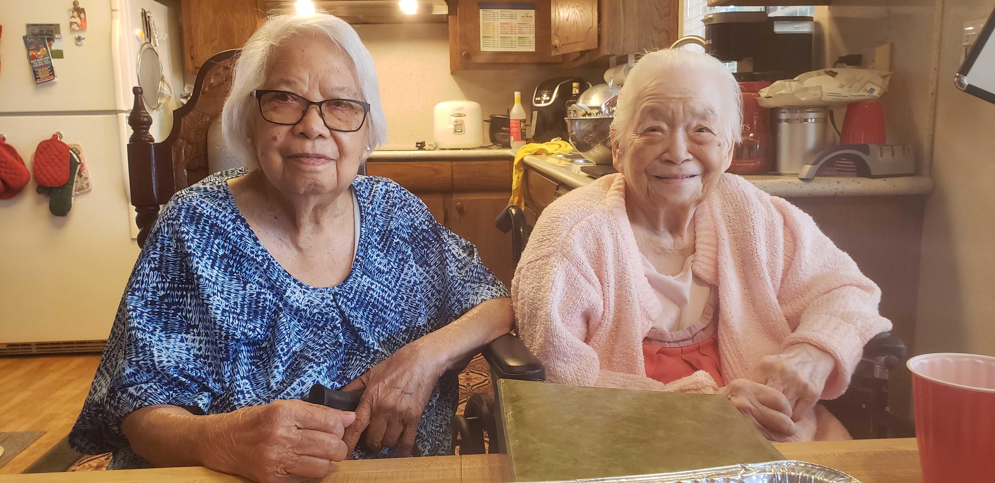 Two elderly women share smiles and conversation at a kitchen table filled with warmth and memories.