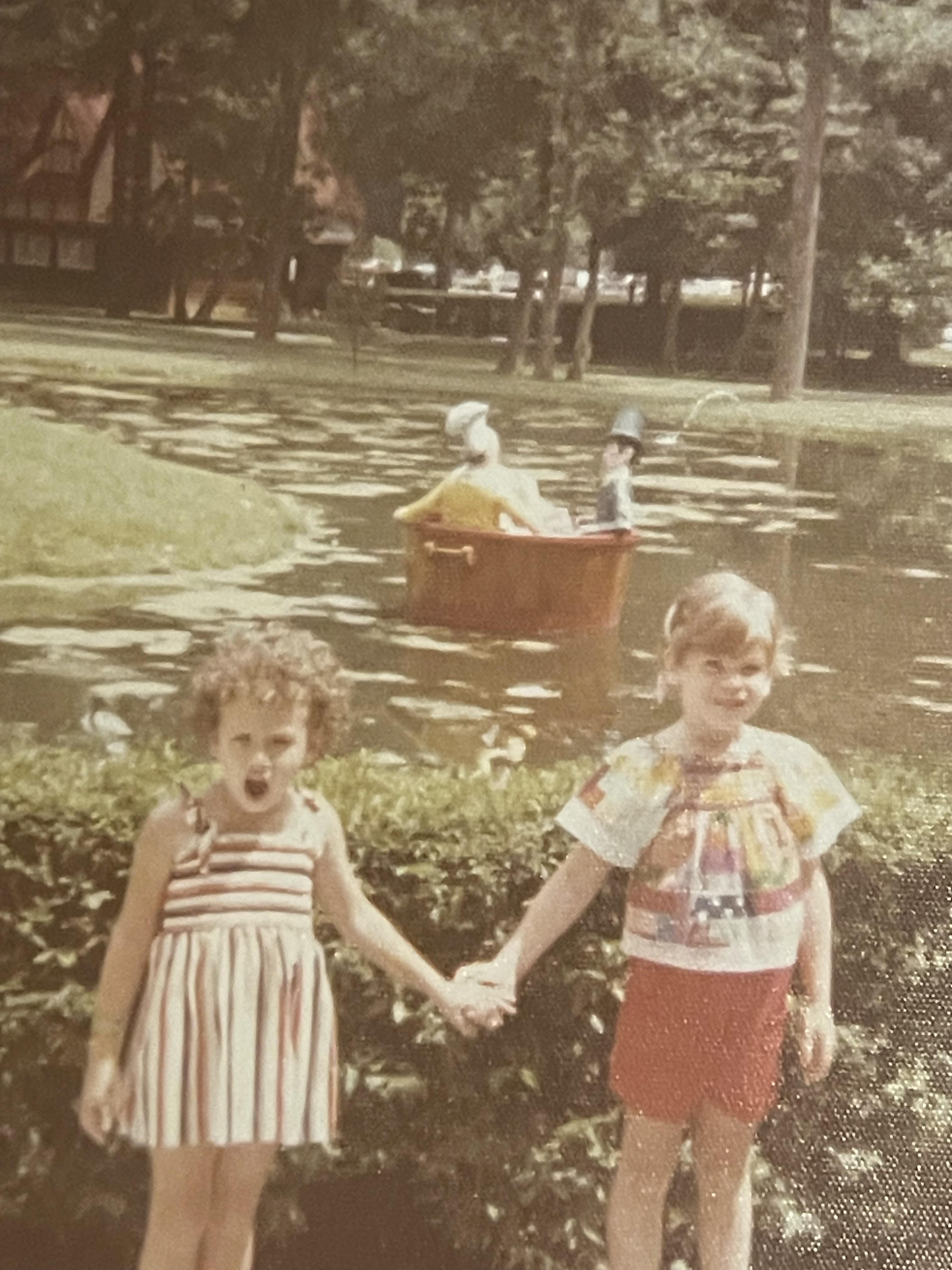 Two girls stand side by side by a pond, smiling as they hold hands while people row in a boat.