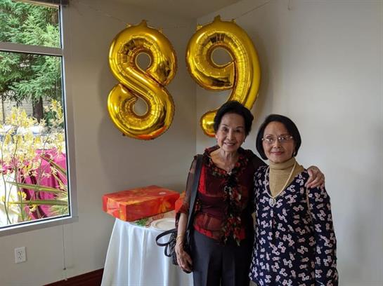 Two women pose happily together at a birthday celebration with large balloons in the background.