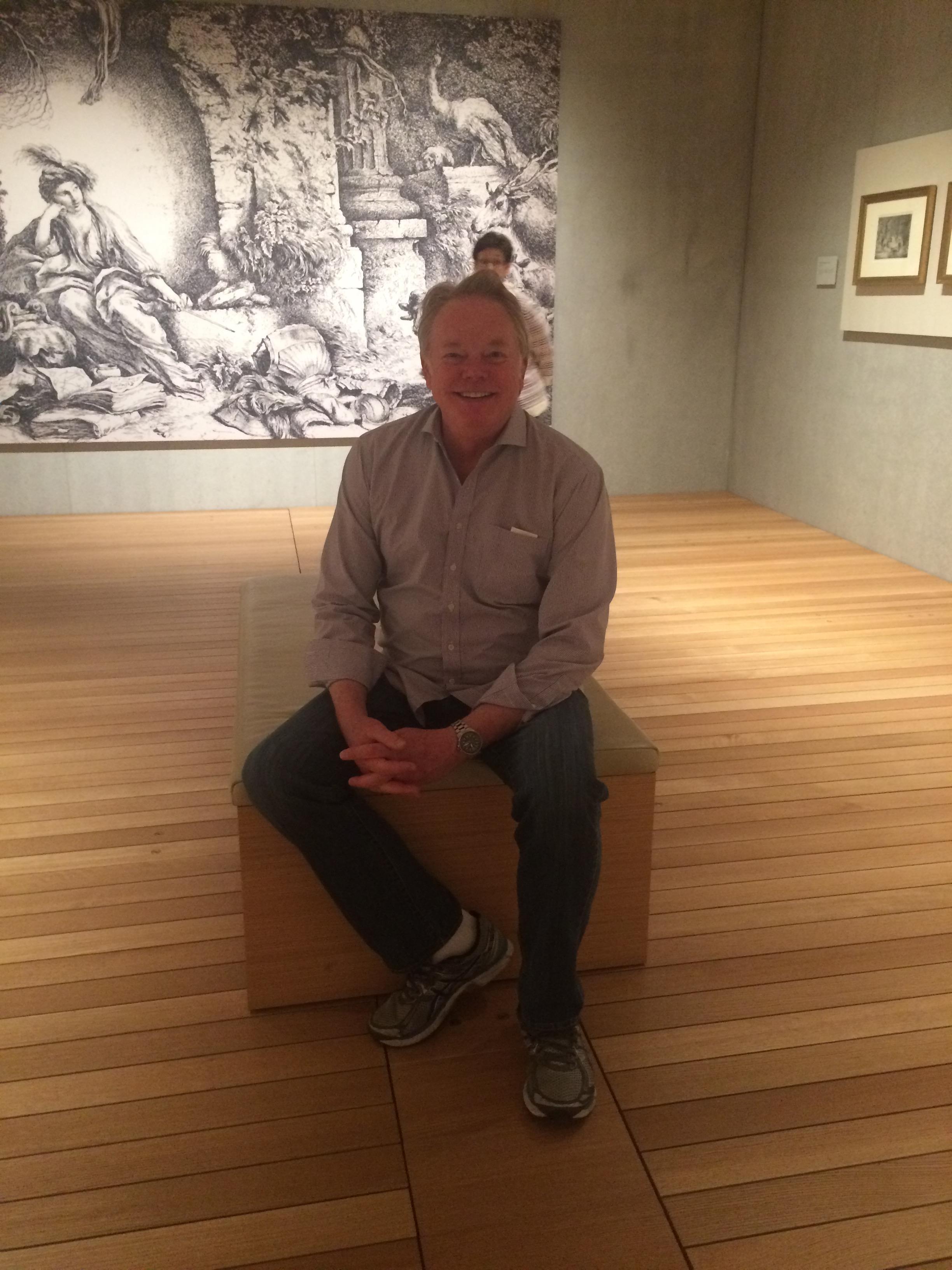 A man sits relaxed on a wooden bench in a contemporary art gallery, admiring nearby artwork.