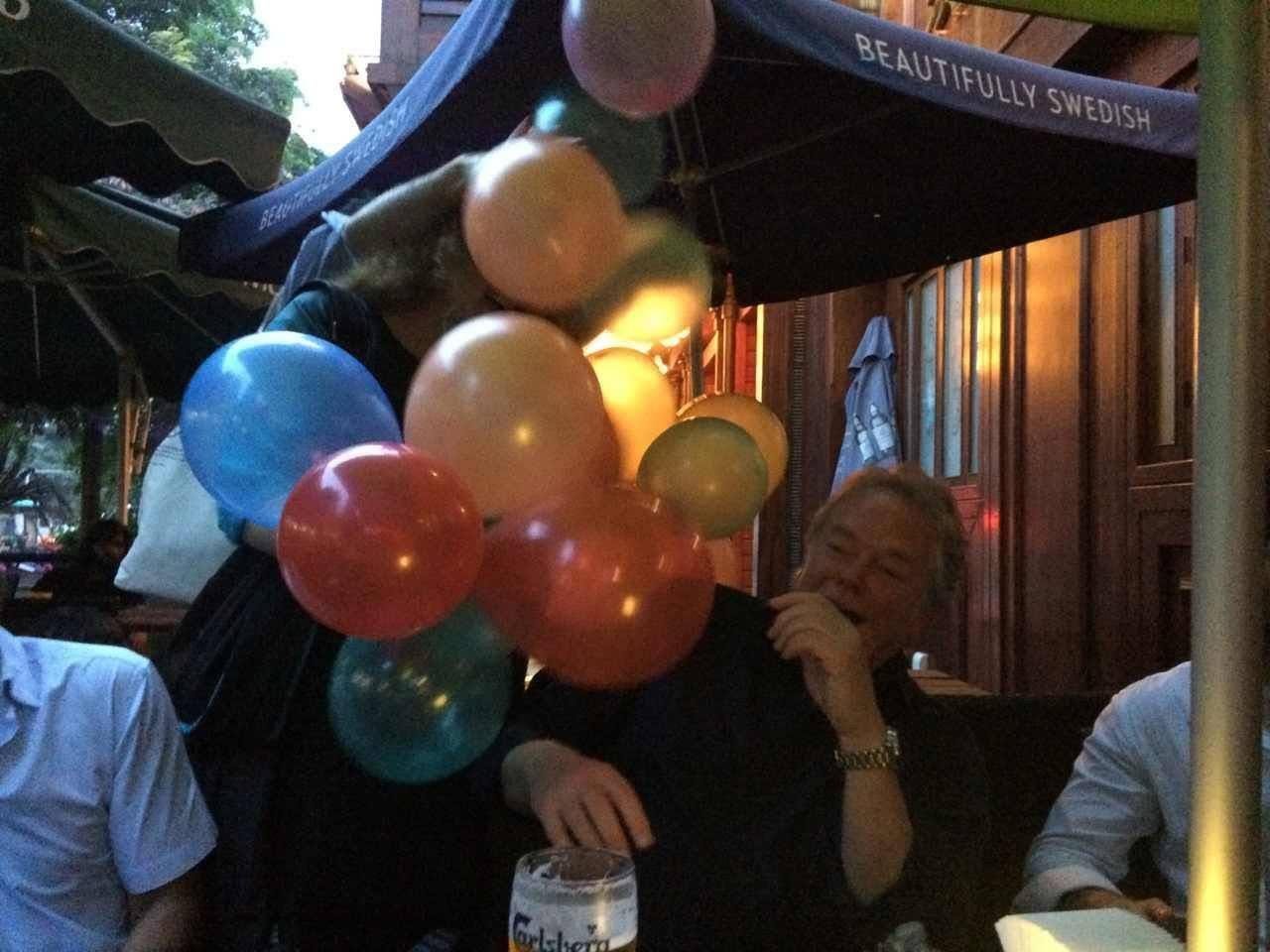 Guests enjoy drinks while a person surprises them with a bunch of colorful balloons at dusk.
