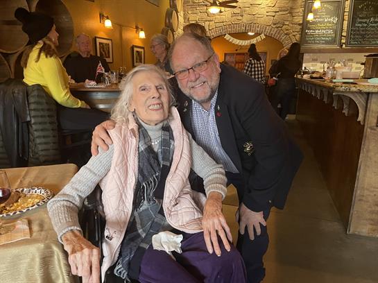 Elderly woman smiles warmly while posing for a picture with a man in a cafe during daytime.