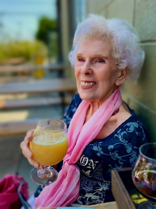 A cheerful woman with white hair holds a colorful drink while seated outside, smiling brightly.
