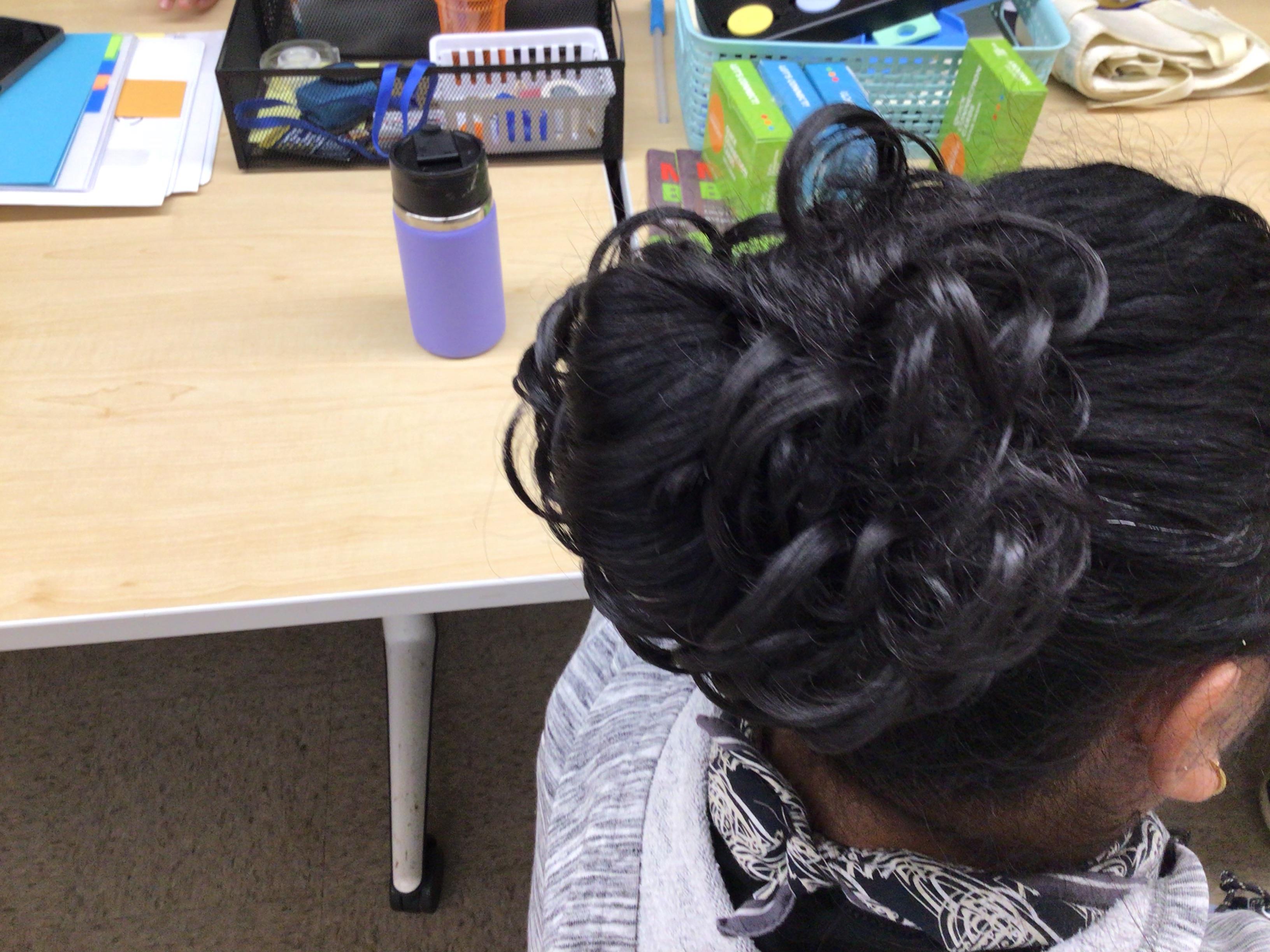 A woman with intricately styled hair is working at a desk filled with colorful classroom supplies.
