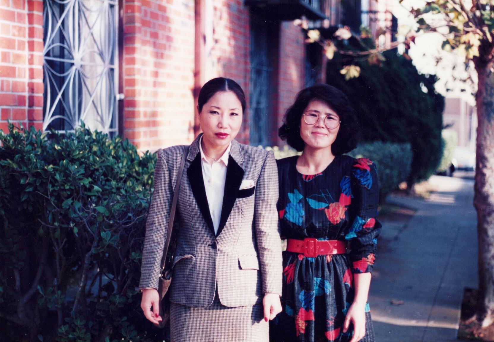 The women stand confidently on a city sidewalk with trees and brick buildings in the background.