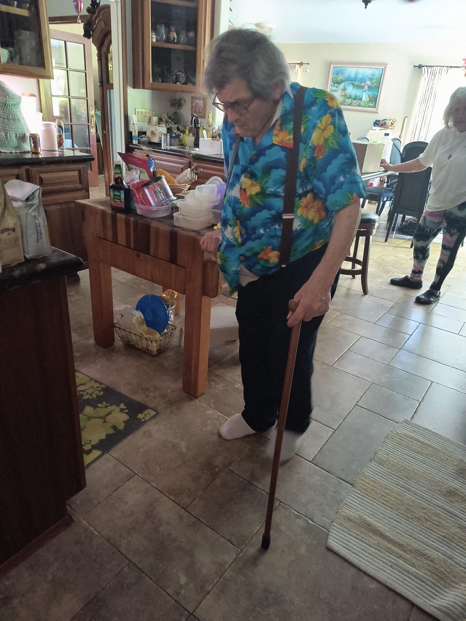 An elderly woman wearing a colorful shirt walks carefully with a cane in a spacious kitchen.