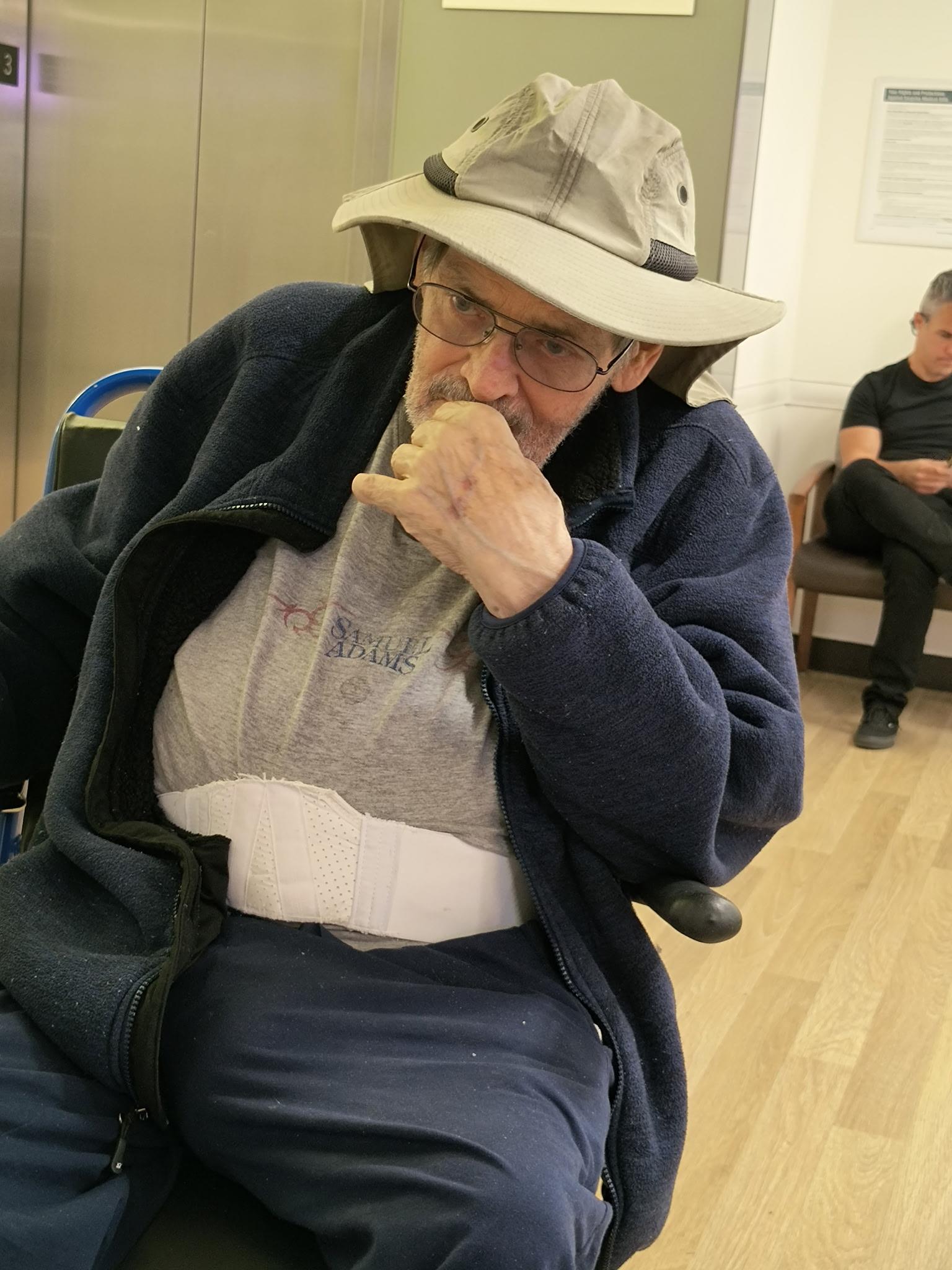 An elderly man in a hat appears contemplative while seated in a waiting area.