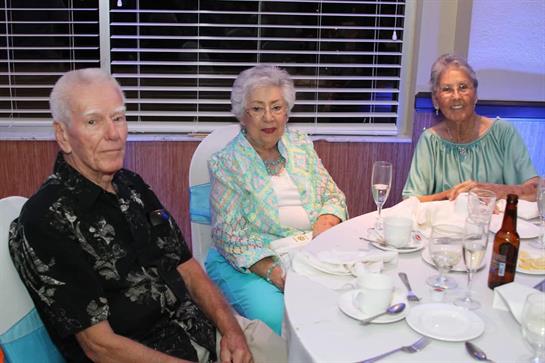 Three elderly individuals engaged in conversation at a festive dinner gathering.