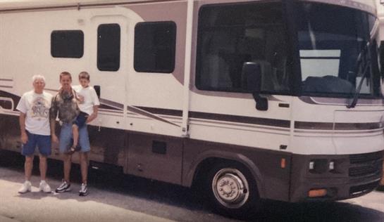 A family poses beside their RV at a campsite under the bright summer sun.