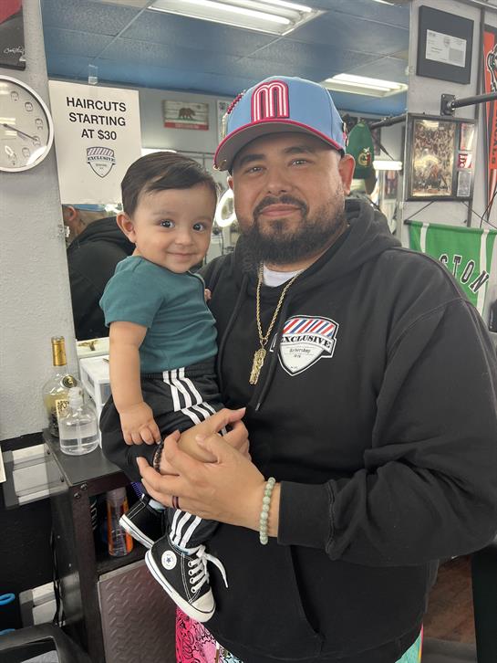 A joyful father holds his young son in a bustling barbershop as they enjoy their day together.