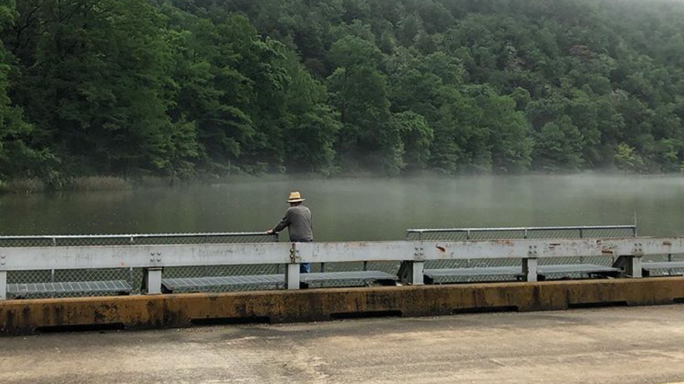A person sits quietly on a bridge casting a fishing line into a misty river surrounded by trees.