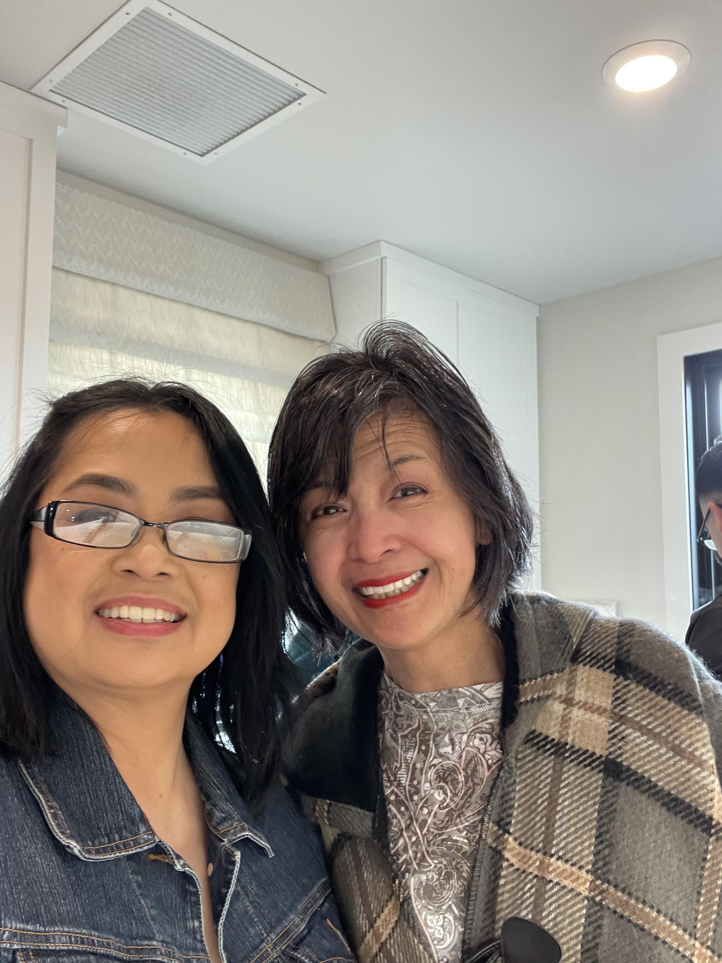 Two friends smile happily while posing near a window in a well-lit room together.
