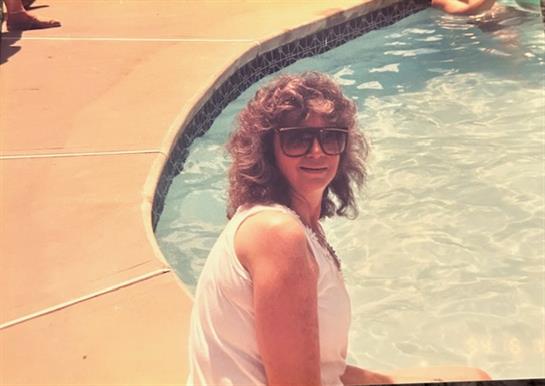 A woman wearing sunglasses sits beside a pool, enjoying the sun and cheerful atmosphere with others.