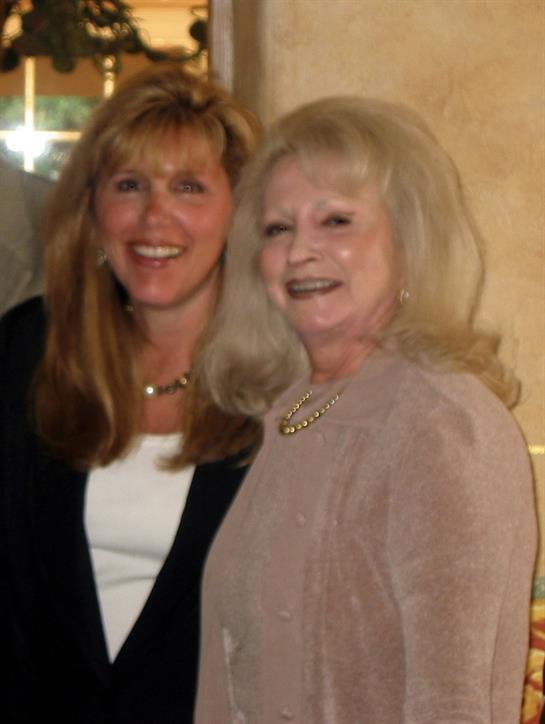 Two women stand close, sharing smiles at a cheerful social event in a cozy environment.