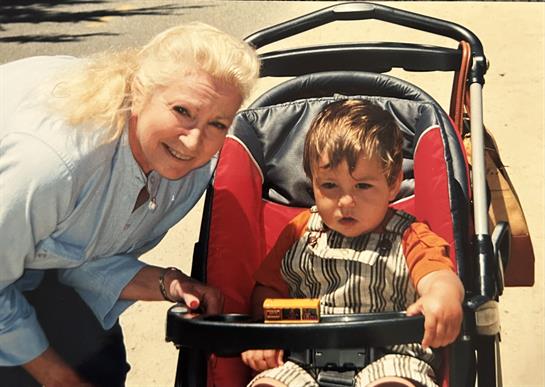 A grandmother joyfully poses beside her grandson in a stroller on a bright day outdoors.
