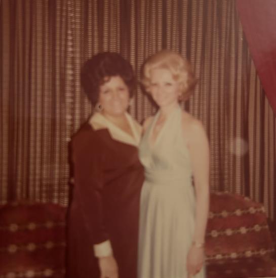 Women are smiling while posing in formal dresses at a gathering with vintage decor.