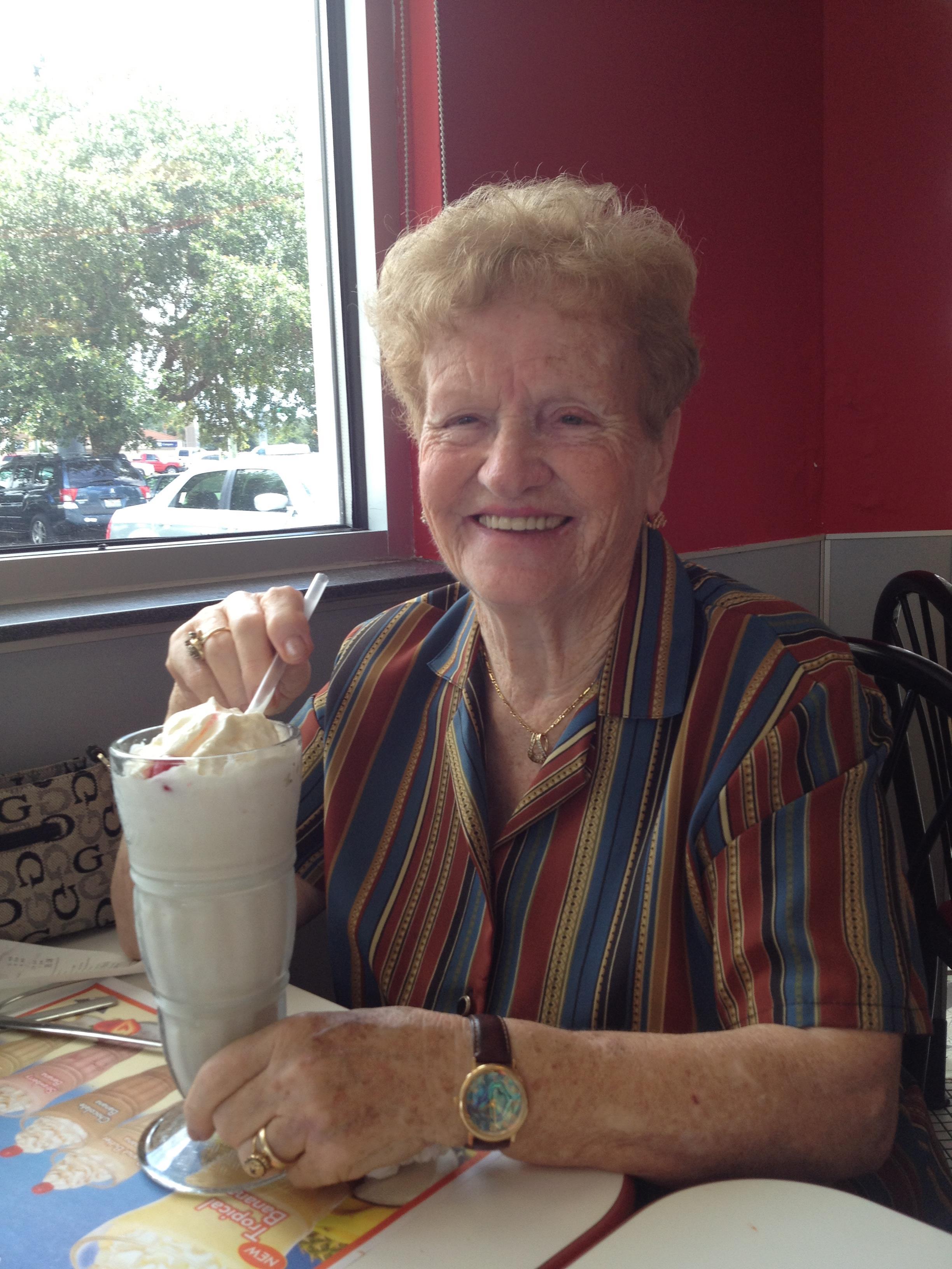 An elderly woman sits at a table, happily holding a large milkshake while smiling warmly.