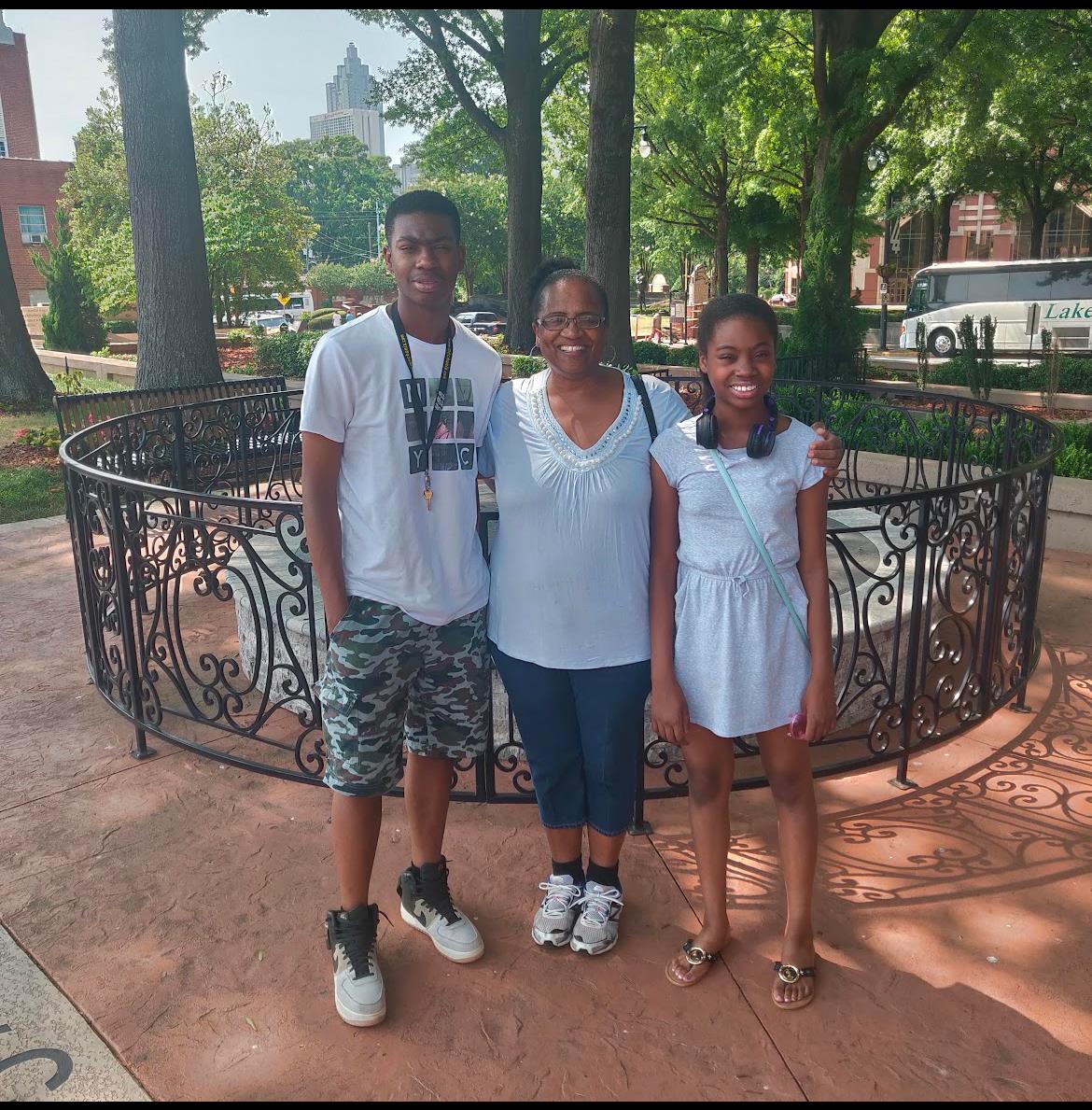 Three family members smile for a picture together, surrounded by greenery in a sunny park.