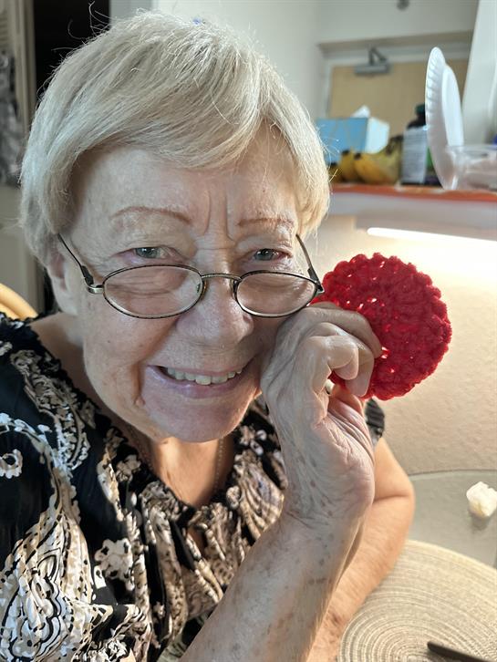 An elderly woman is smiling and holding up a red crocheted piece in a cozy living room.