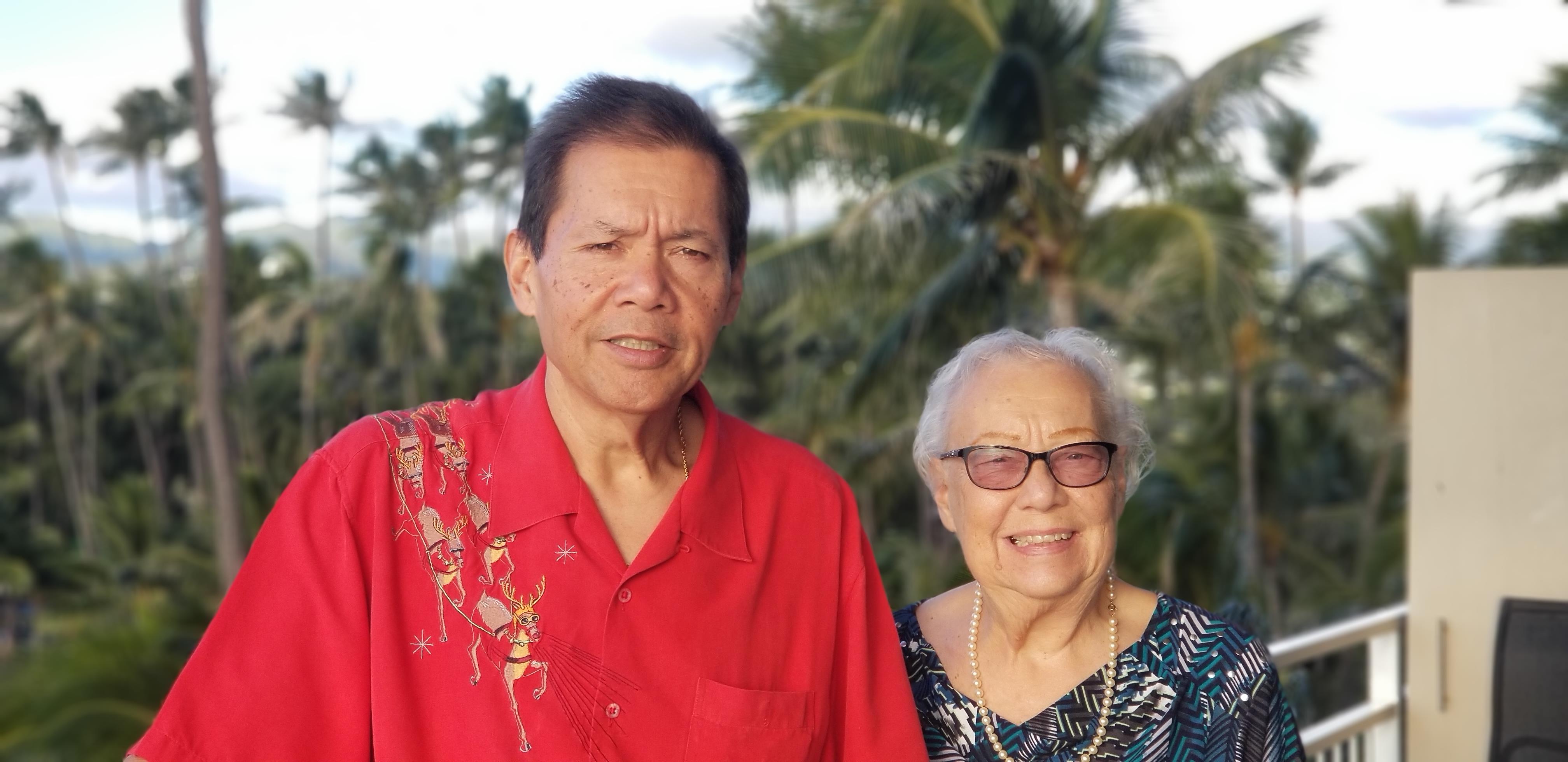 Elderly couple stands together smiling in a vibrant tropical garden under clear skies.