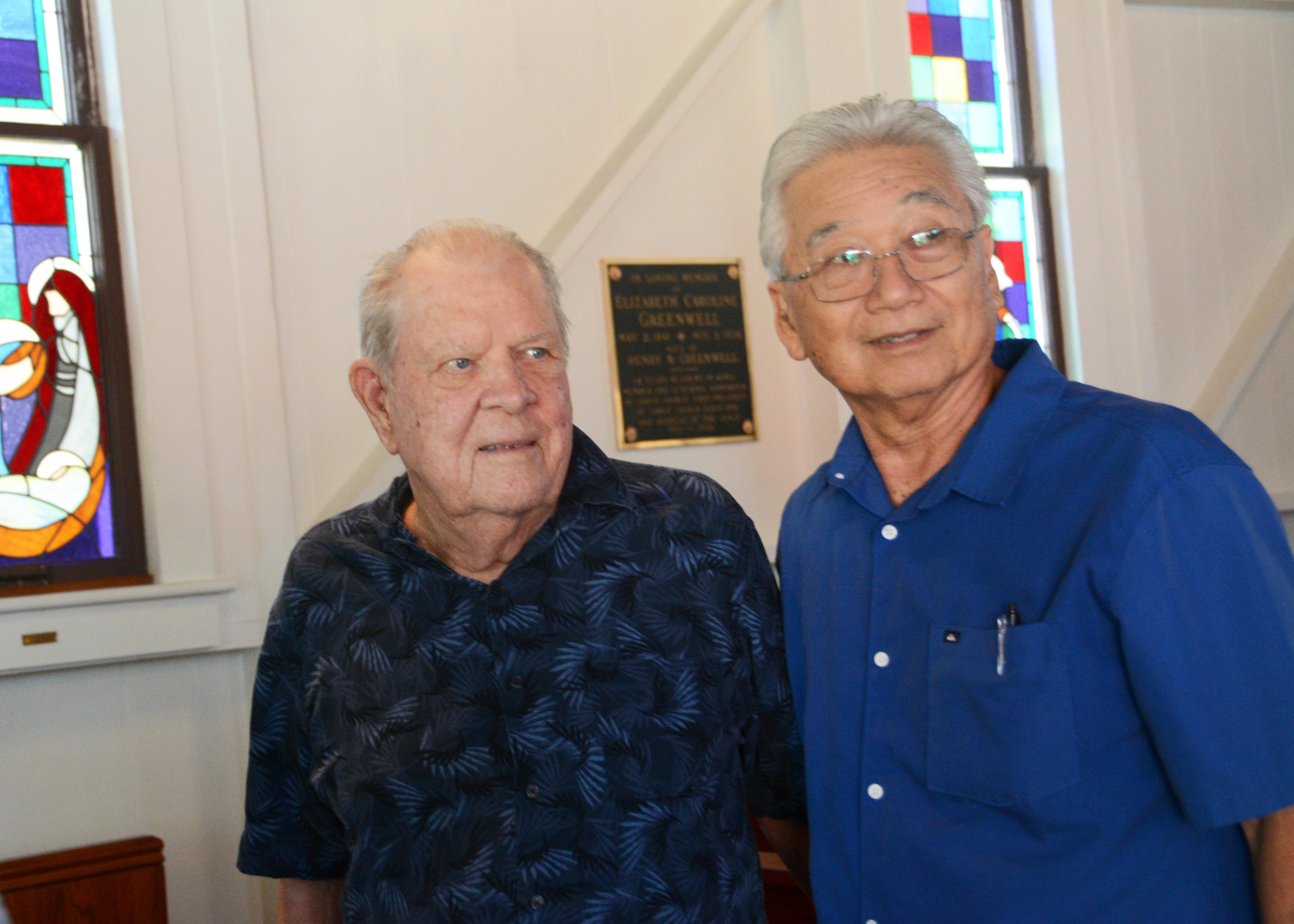 Two older men stand together and smile warmly inside a community center filled with colorful light.